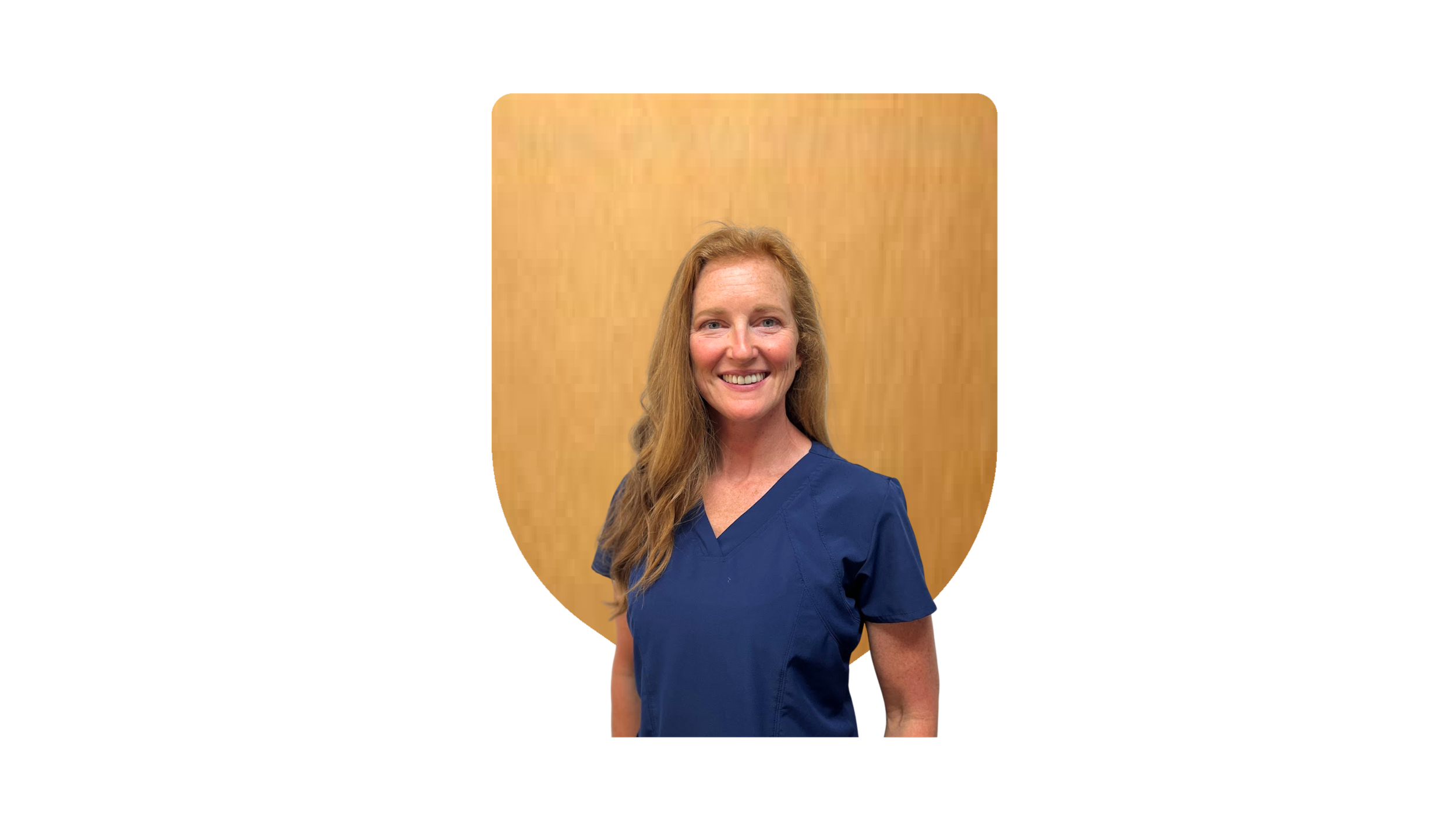 Portrait of a smiling woman with long, wavy red hair wearing a navy medical scrub top, standing in front of a wooden background.