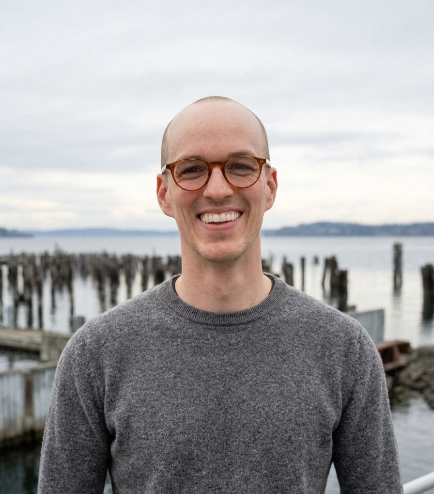 Portrait of a smiling man with glasses standing outdoors near water, with wooden posts and a cloudy sky in the background.