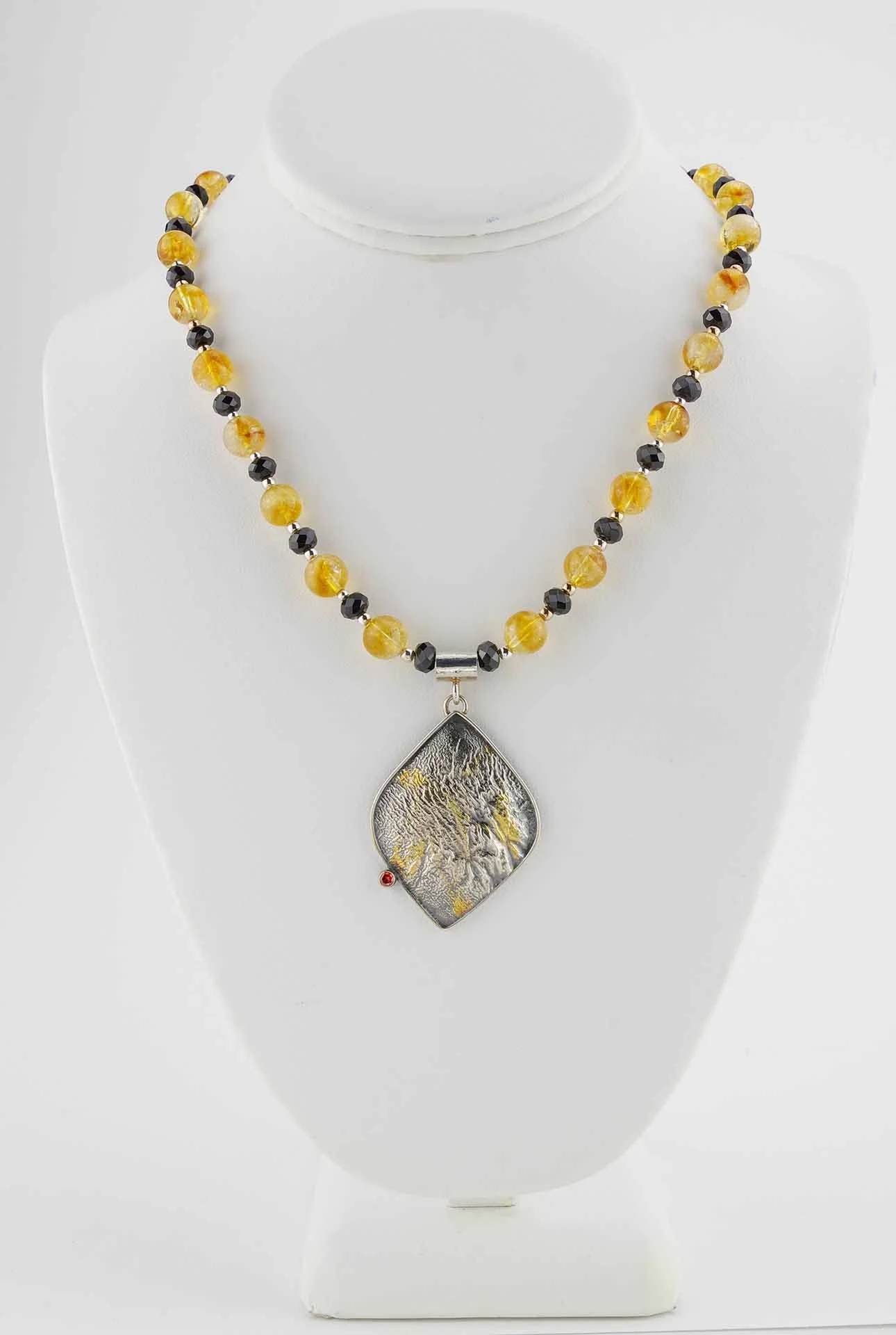 Necklace of citrine beads, faceted black rondelles and silver accents with a large, slightly irregular shaped oxidized silver pendant. Shown hanging on a jewelry display.