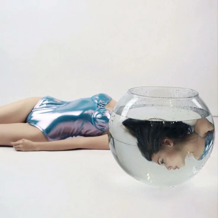 A woman lying on the ground wearing a shiny, metallic dress with a large fishbowl of water placed in front of her, reflecting her upside-down face.