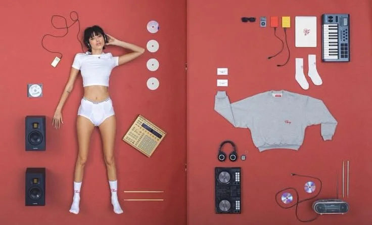 A woman lying on a red floor surrounded by music equipment, clothes, and accessories.