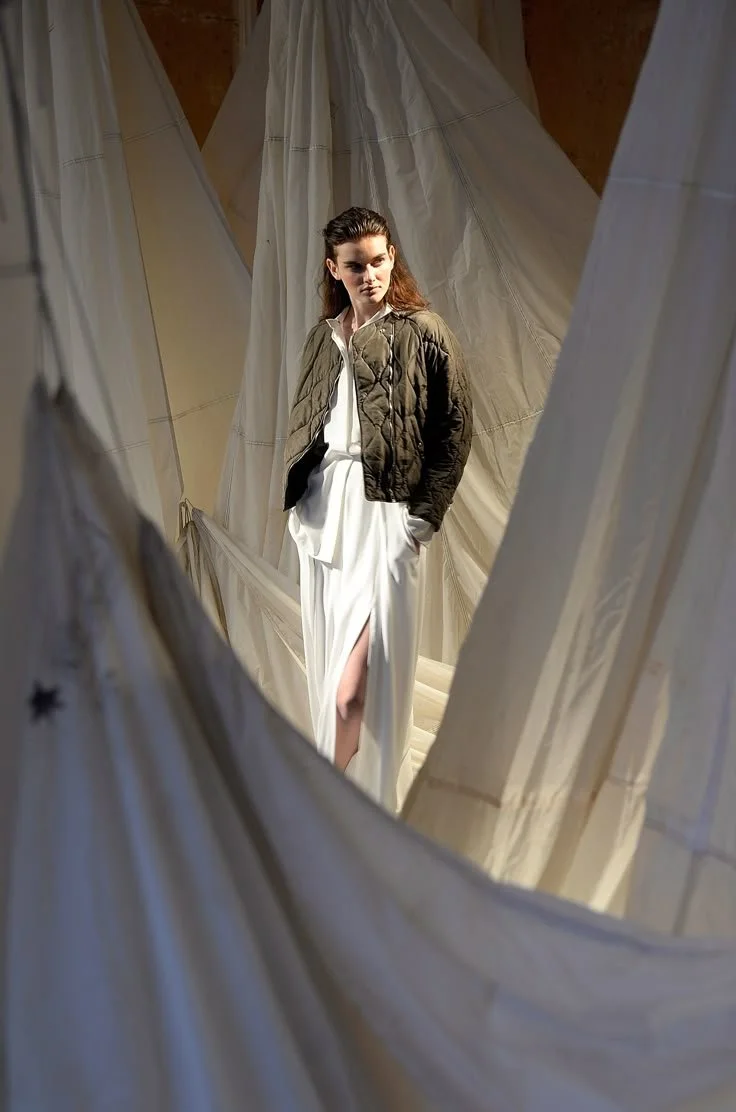 A woman standing among hanging fabric drapes, wearing a white dress and a brown jacket.