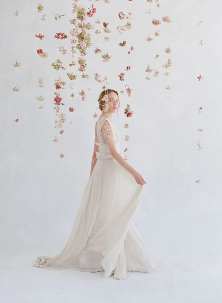 A woman in a white wedding dress holding the edge of her gown, standing against a backdrop with hanging flowers and butterflies.