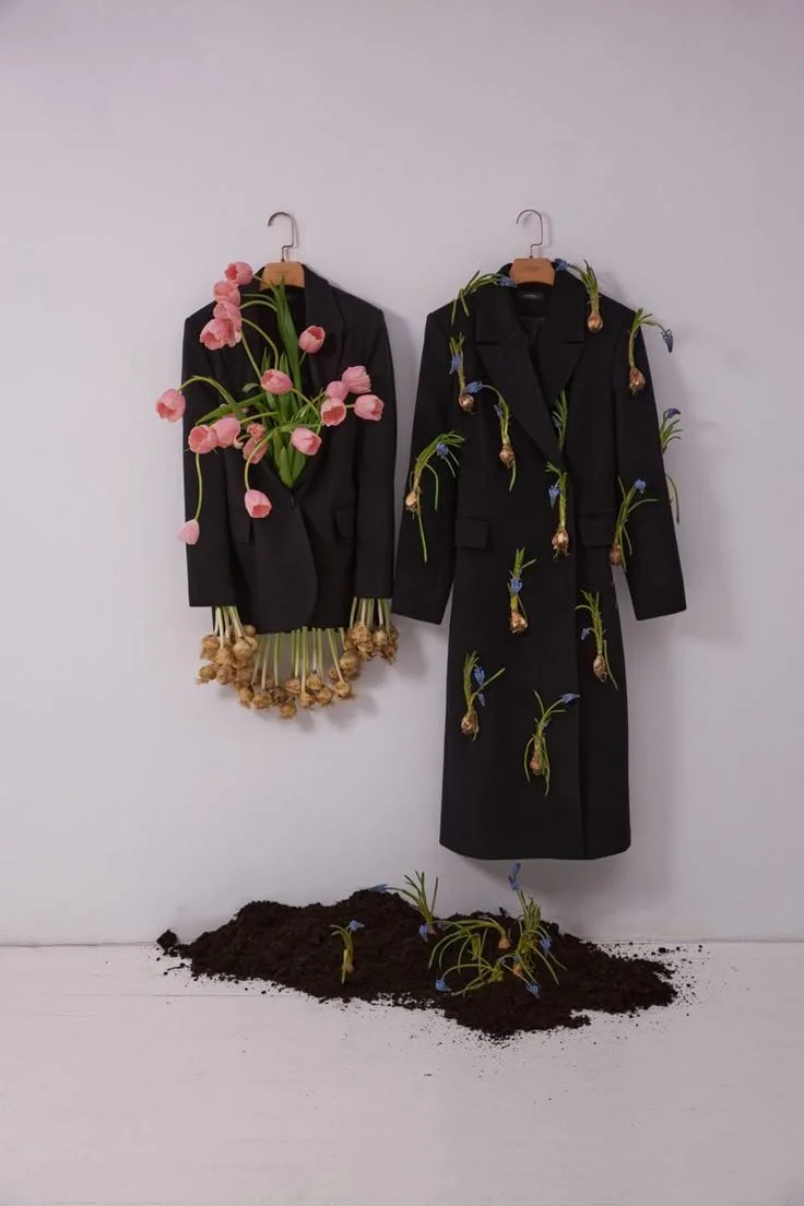 Two black suits decorated with flowers and bulbs, hanging on a plain white wall above a patch of soil with more flowers growing from it.