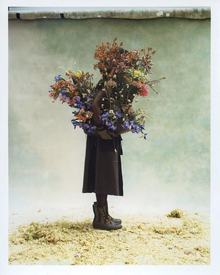 Person holding a large bouquet of colorful flowers, standing against a plain background, wearing a black coat and boots.