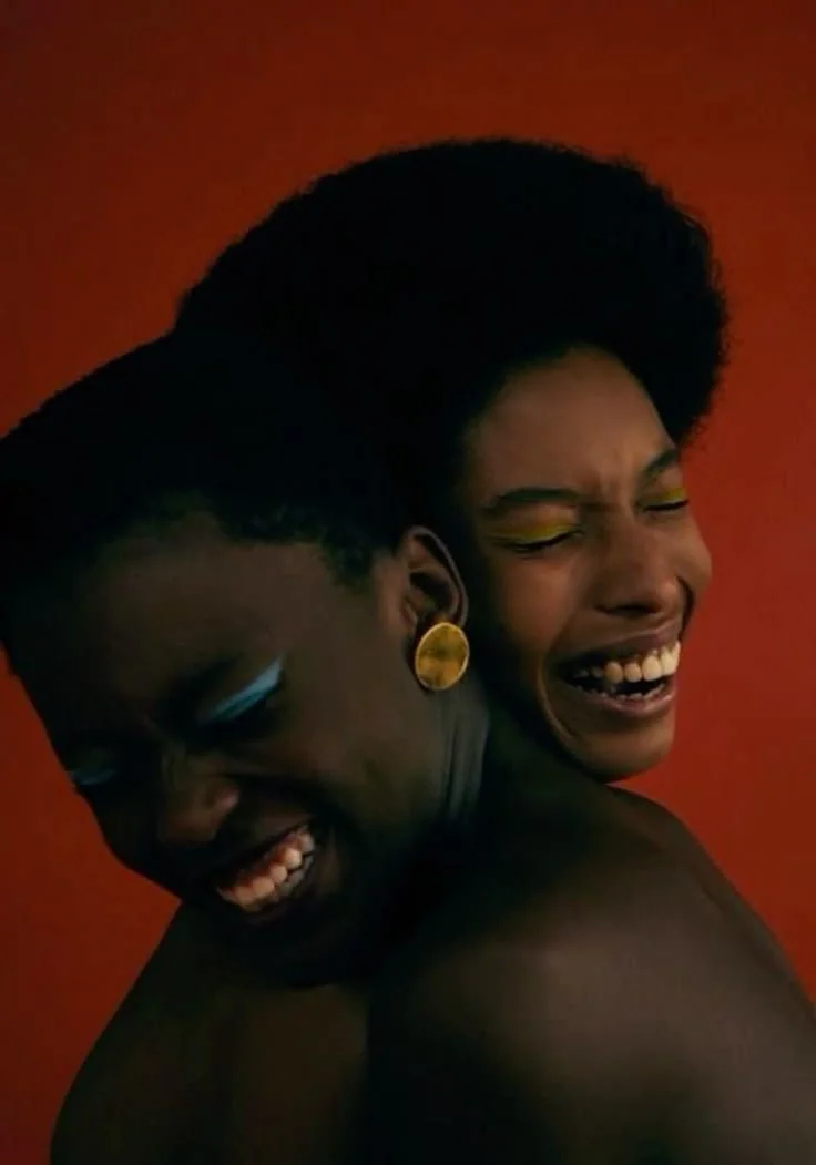 Two women with dark skin, laughing and hugging each other, against a red background, one with short hair and gold hoop earrings, the other with an afro hairstyle and yellow eye makeup.