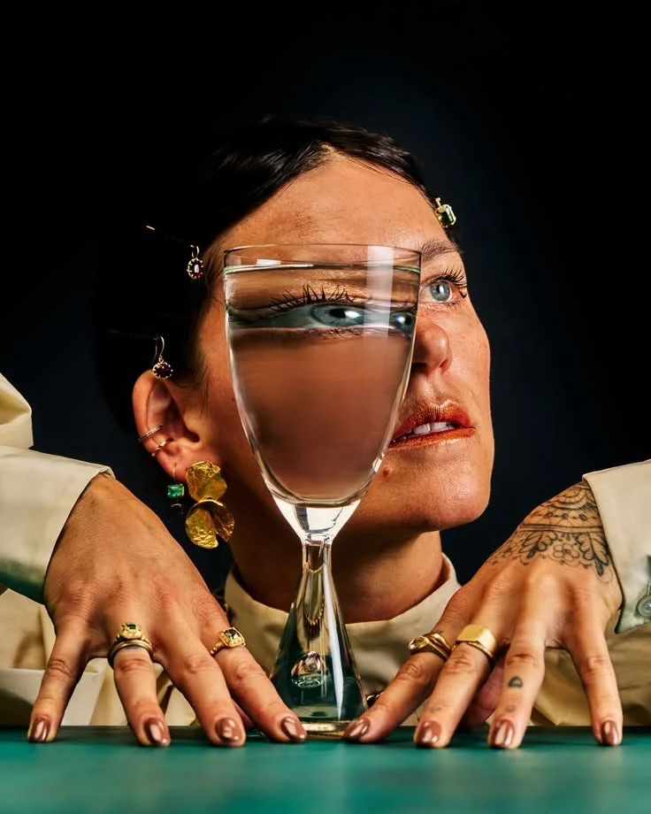 A woman with tattoos and jewelry resting her hands on a table with a large glass of water in front of her, reflecting her eyes.