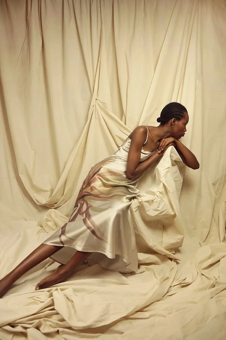 A woman in a satin dress is posed on a draped cream-colored fabric background, leaning on a chair or prop, with one arm bent and her head resting on her hand.