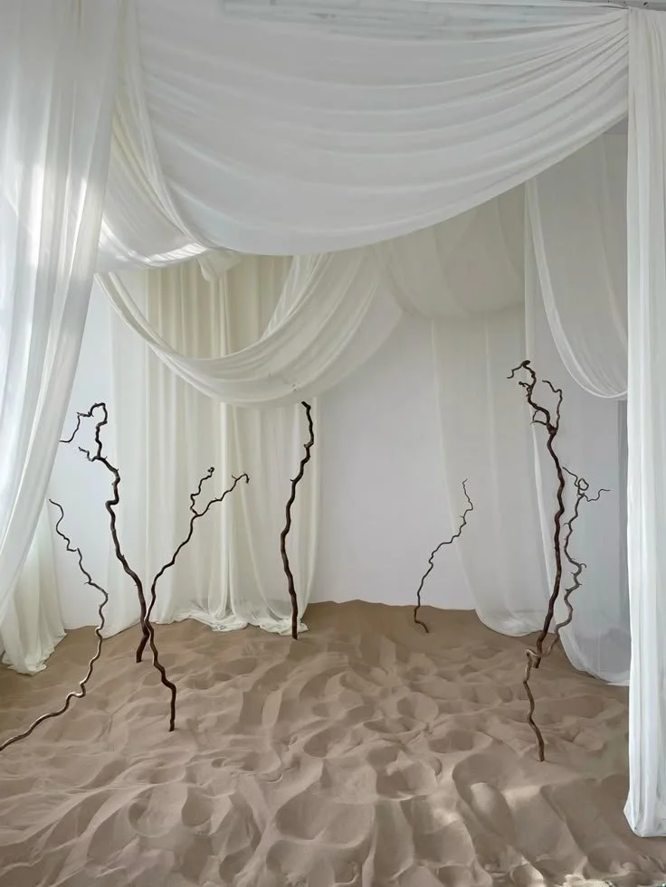 Interior of a room with white draped fabric walls and ceiling, and sand on the floor, with several twisted, leafless branches standing upright.