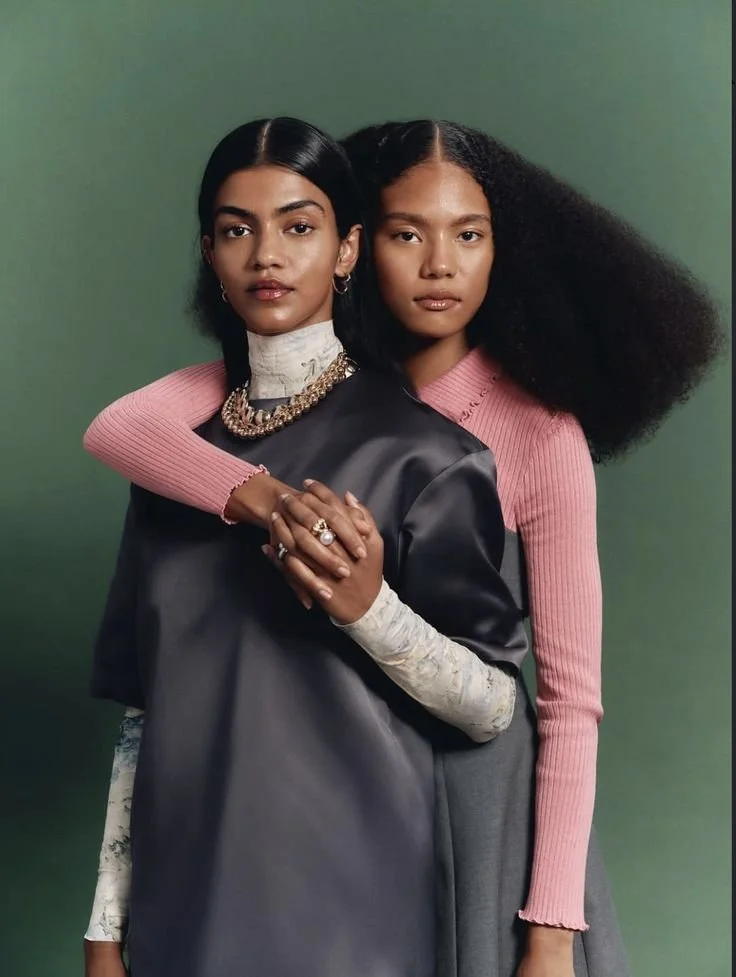 Two women standing closely together, one with straight black hair and the other with curly hair, against a green background. The woman with straight hair is wearing layered necklaces and a black satin jacket, while the woman with curly hair is wearin