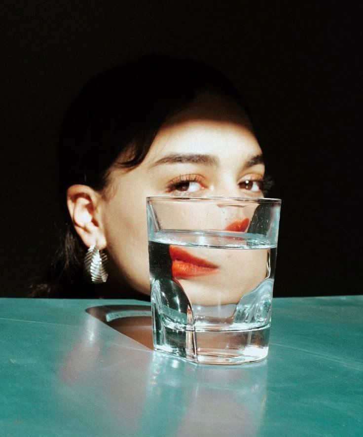 A woman with dark hair wearing earrings and red lipstick, partially obscured by a glass of water on a teal surface, with her face reflected in the glass.