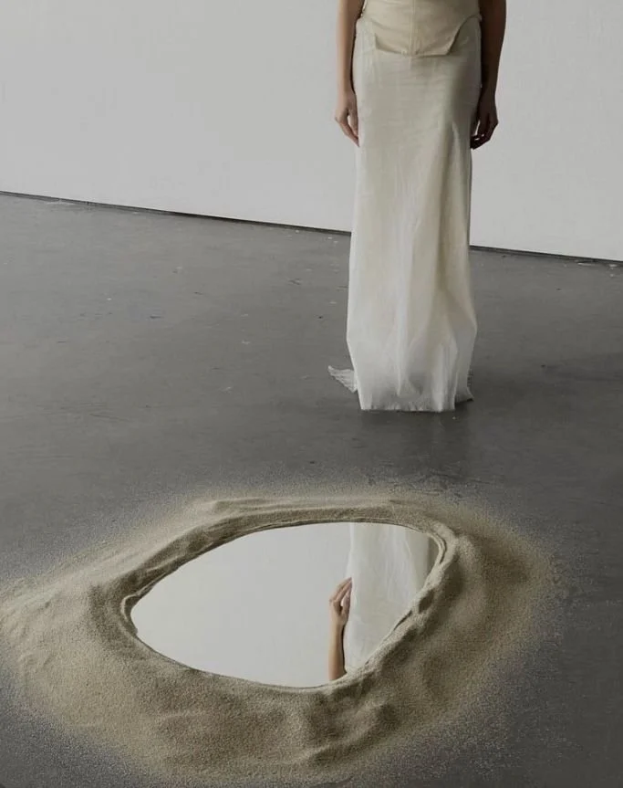 A person in a long cream-colored dress stands next to a hole in the floor filled with a mirror reflecting their hand and part of arm. The floor is gray and the background is plain white.