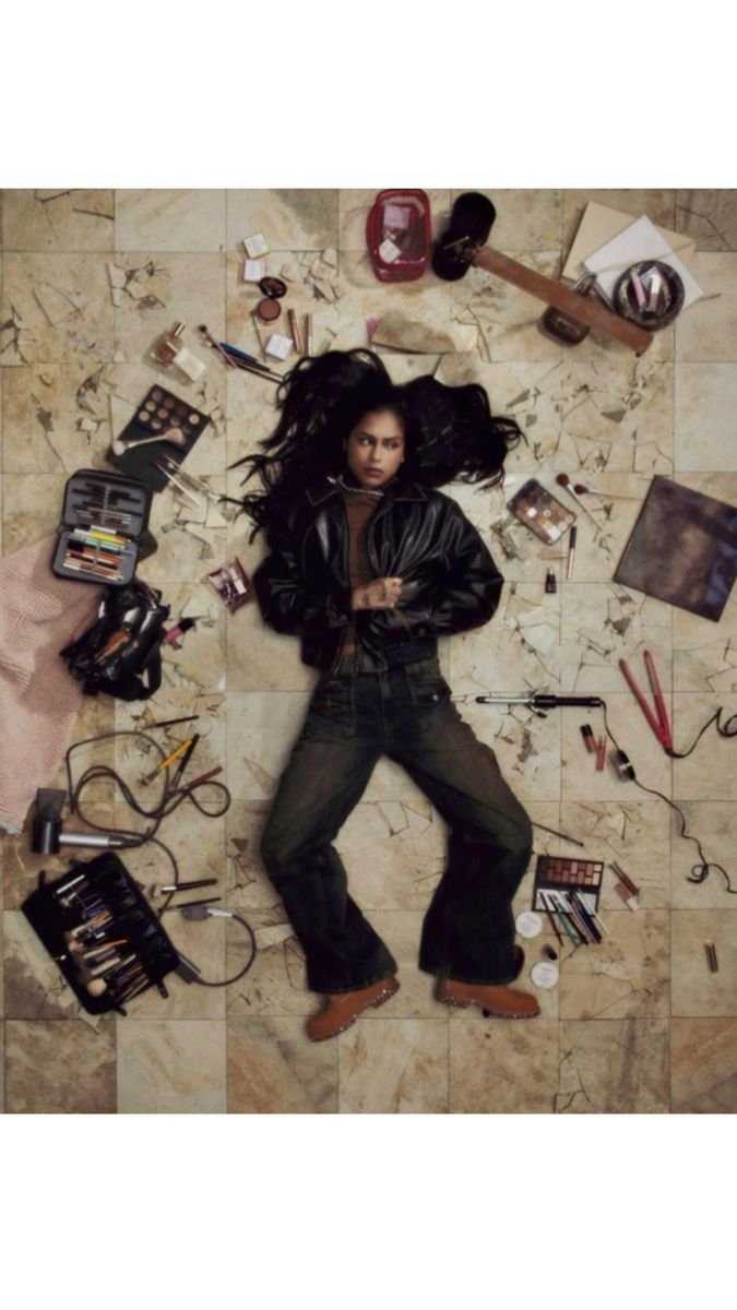 A woman lying on a marble floor surrounded by makeup tools and cosmetics, wearing a black leather jacket and brown boots.
