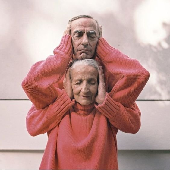 An elderly man and woman are standing close, both covering their ears with their hands and having serious expressions.