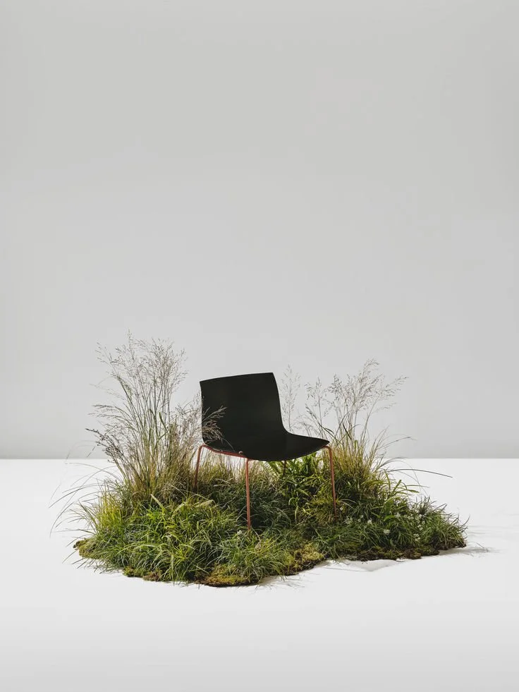 A black modern chair is placed on a small patch of grass with tall plants, set against a plain light gray background.