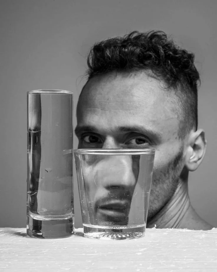 A black and white photograph of a man with a partial view of his face seen through two glasses of water placed on a surface. The man has curly hair and a serious expression.