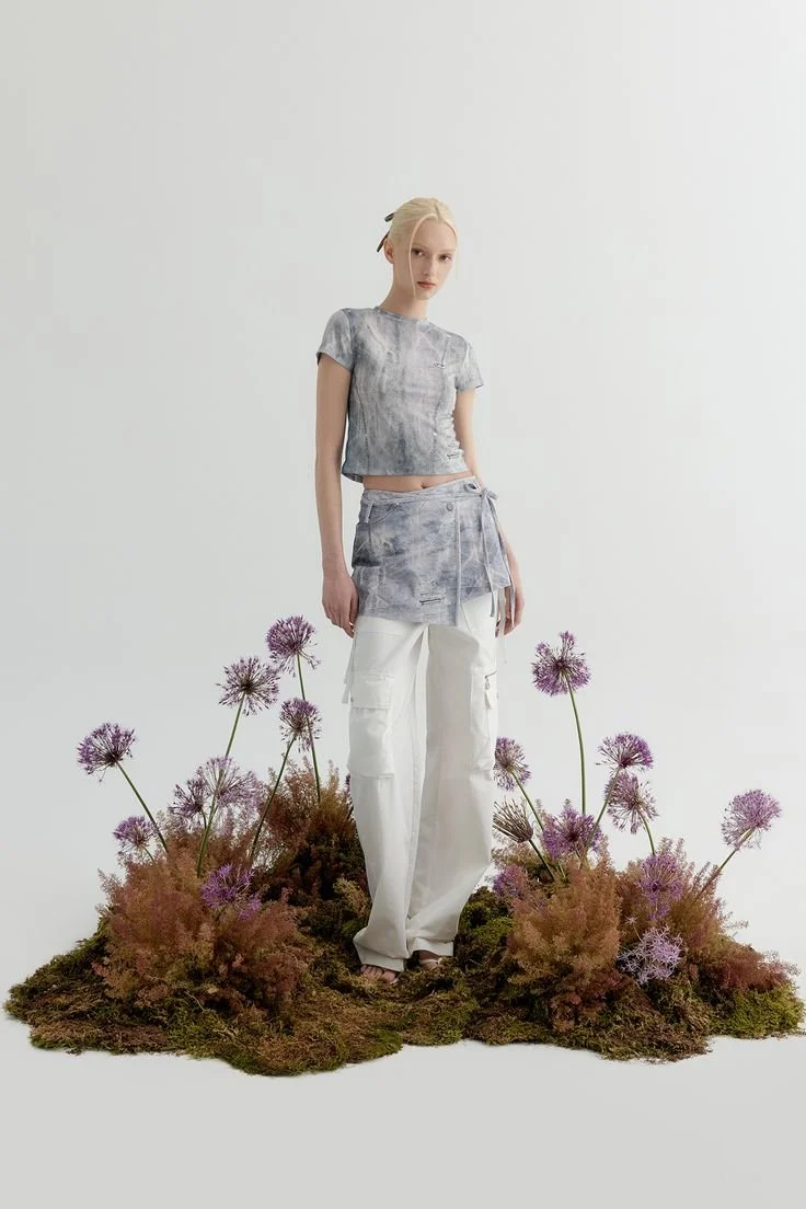 A woman standing on a patch of moss with purple flowers, wearing a gray tie-dye crop top and white cargo pants.