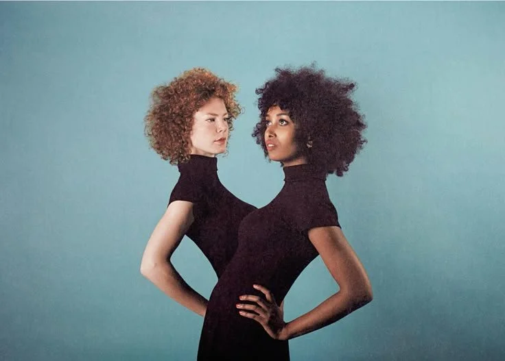 Two women with curly hair in black turtleneck dresses standing close together against a plain background.