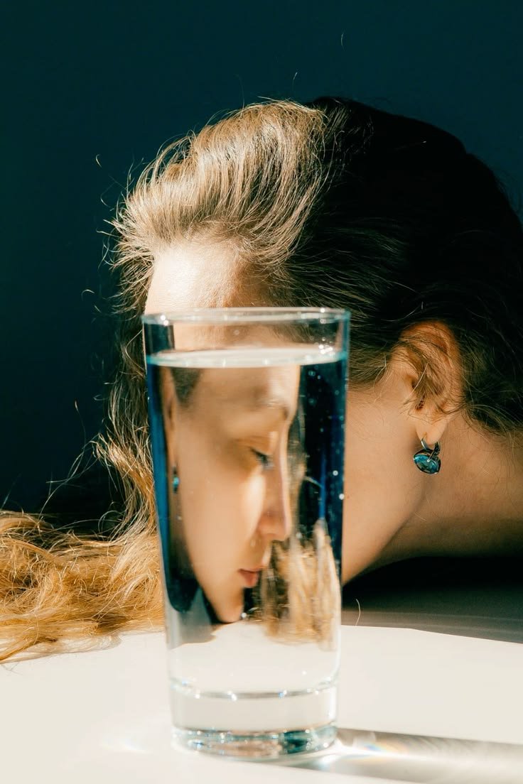 A woman has her face resting on a table, with her eye closed, behind a tall glass of water. The glass reflects her face and hair.