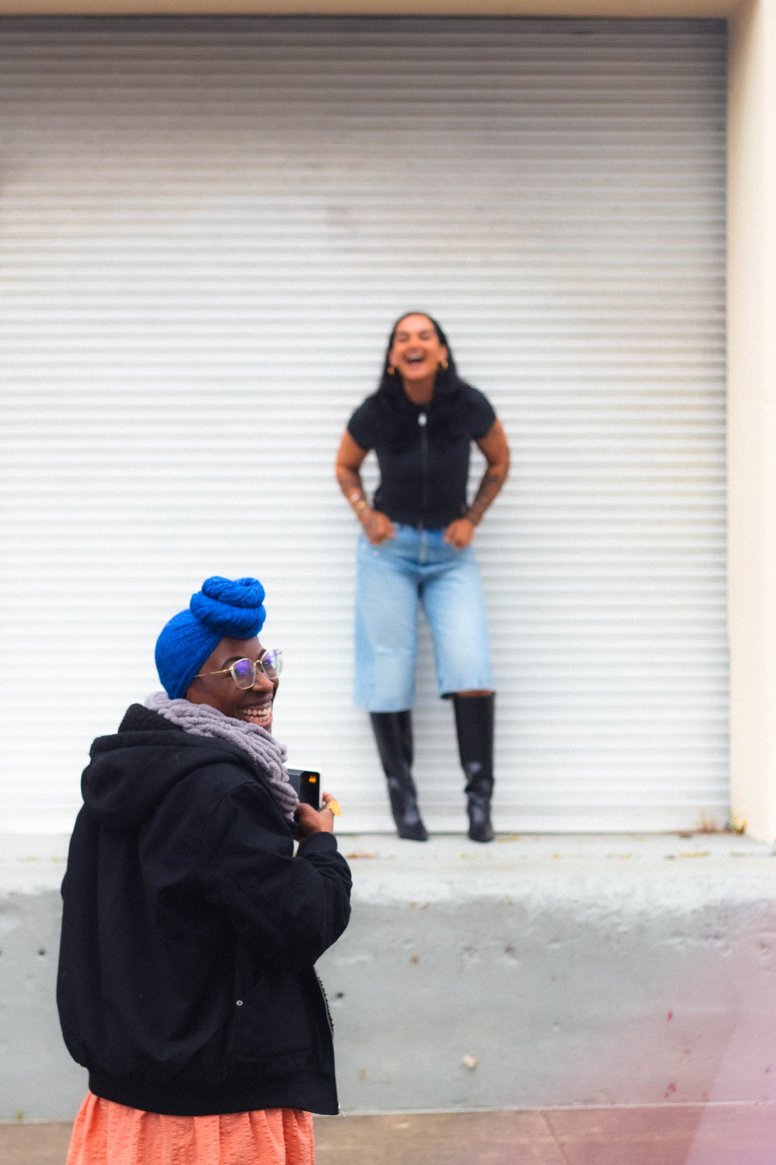 Two women are standing outdoors in front of a large white roller shutter door. The woman in the foreground has dark skin, glasses, and a bright blue head wrap, wearing a black jacket and orange pants, holding a smartphone. The woman in the background has medium skin, long dark hair, wearing a black top, light blue jeans, and black boots, laughing with her hands in her pockets.