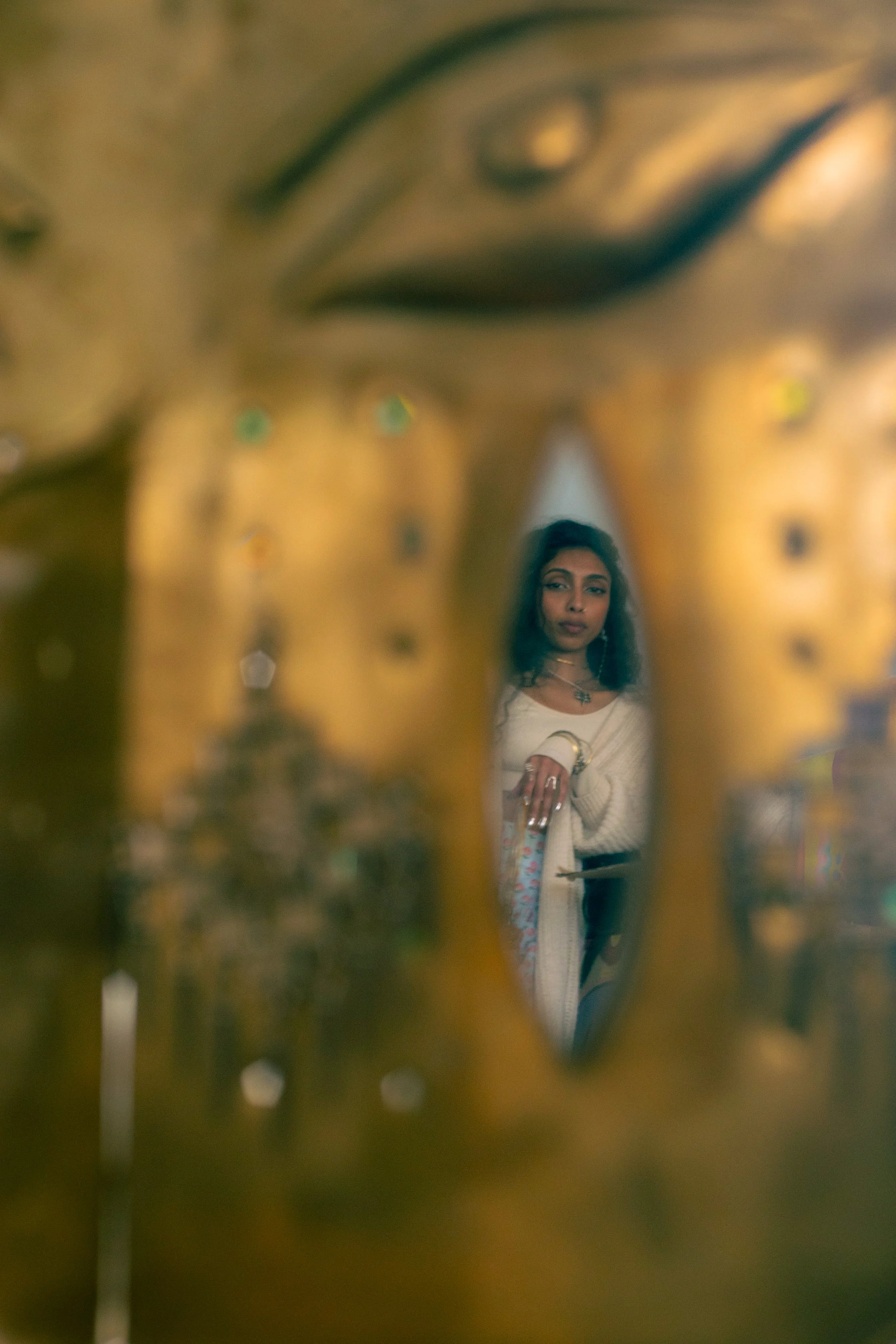 A woman with dark hair, wearing a white sweater and jewelry, is looking at the camera through a small, heart-shaped opening in a golden, ornate decorative surface.