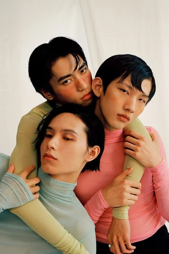 Three young adults in pastel long-sleeve shirts pose closely together, with two of them embracing the third, against a plain white background.