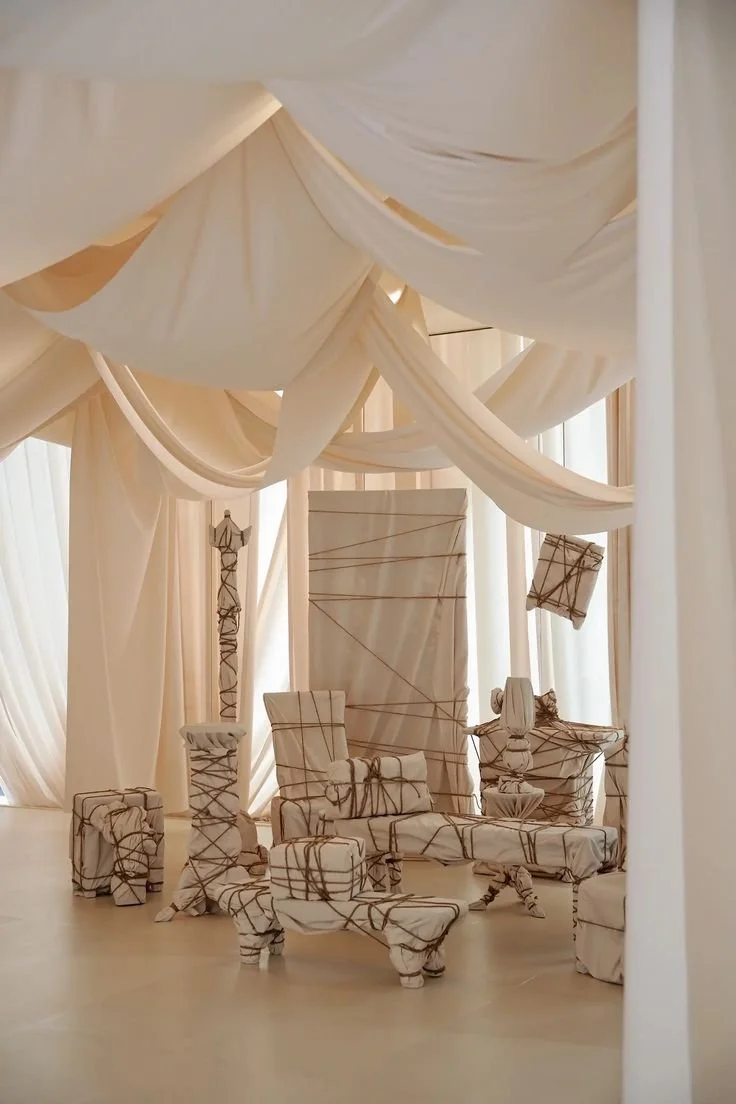 Room decorated with cream-colored drapes and furniture wrapped in beige fabric with brown string ties, resembling a wrapped gift or sculpture installation.