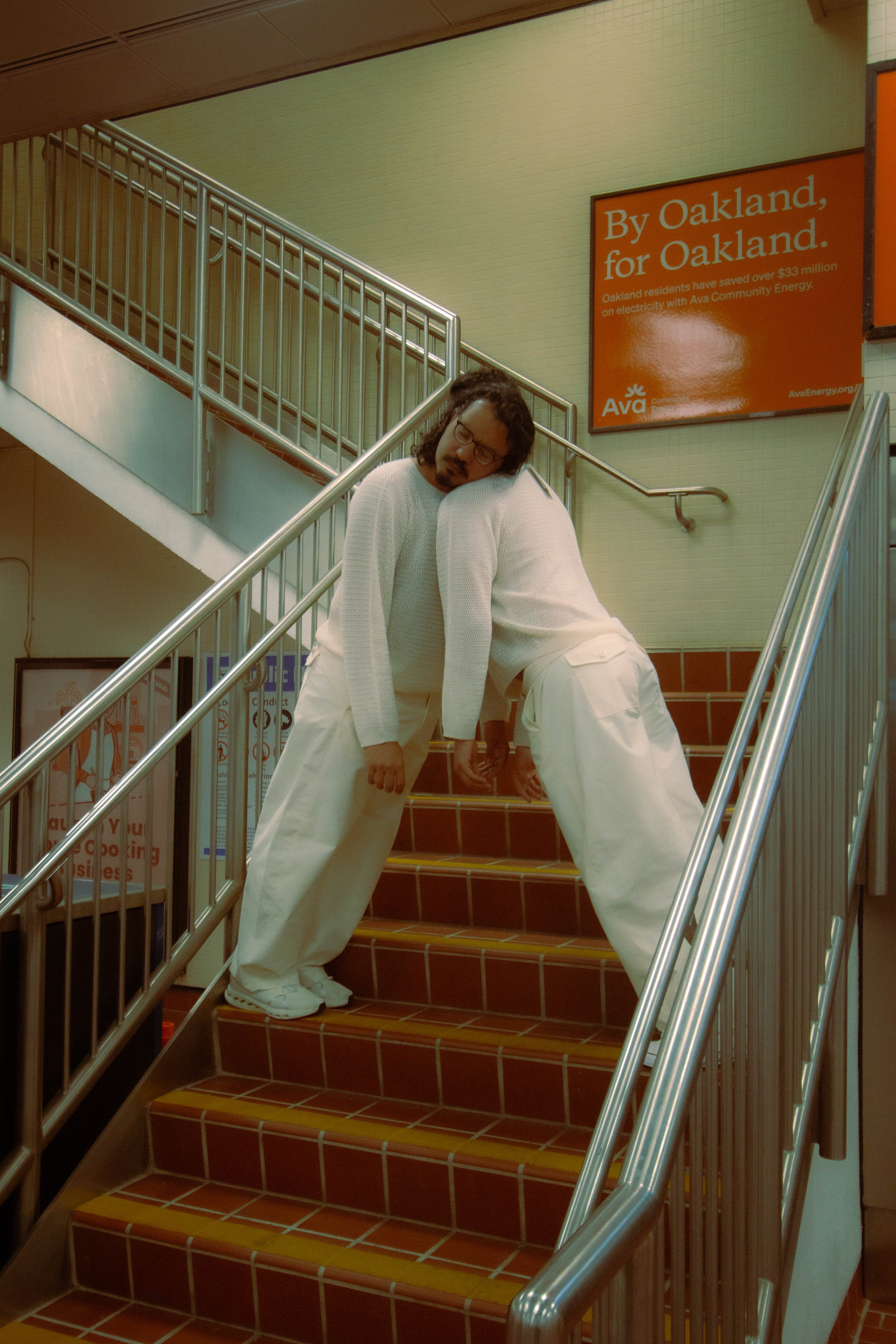 Two men with curly hair and glasses are posing on a tiled staircase with a metal handrail, in front of an orange sign that promotes energy savings for Oakland residents.