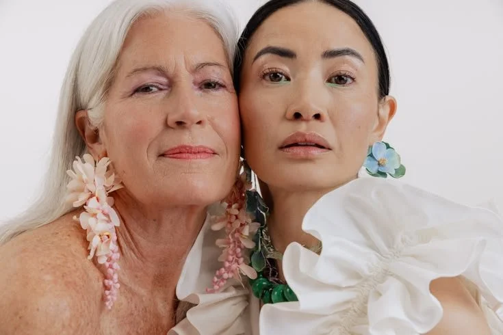 Close-up of two women, an elderly woman with long white hair and a younger woman with dark hair, both wearing floral earrings and white clothing, standing close together against a white background.