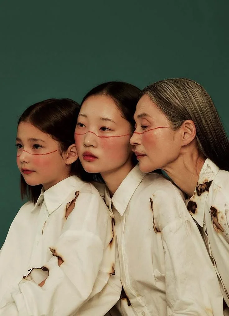 Three females with burnt marks on their faces and torn white shirts standing close together against a green background.