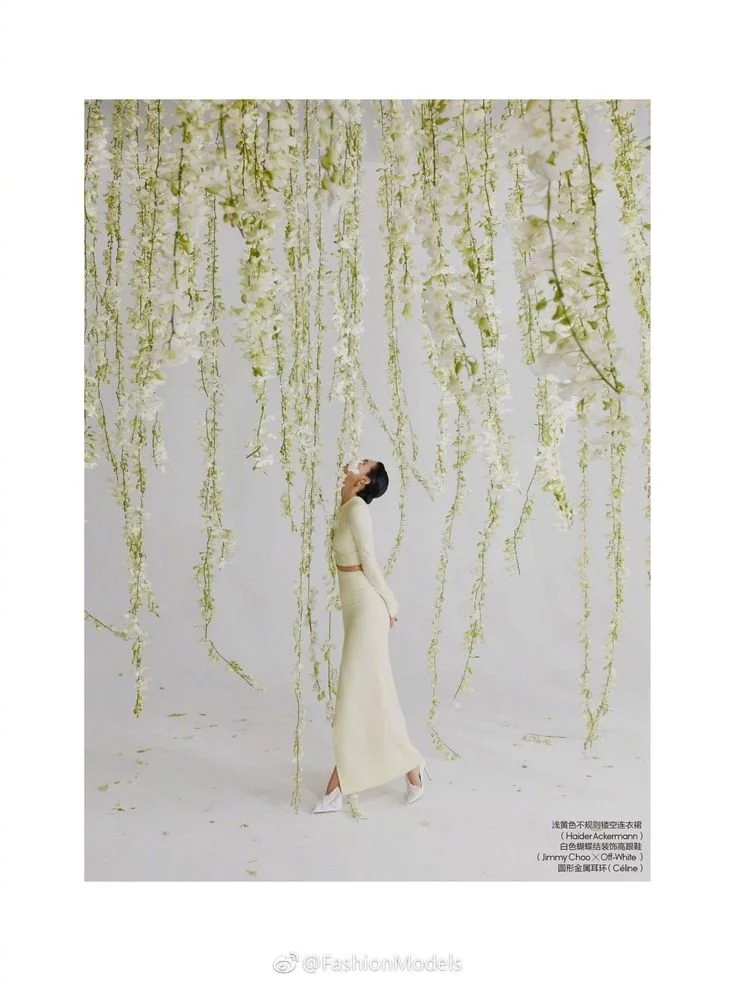 A woman in a white dress standing beneath hanging white flowers against a white background.