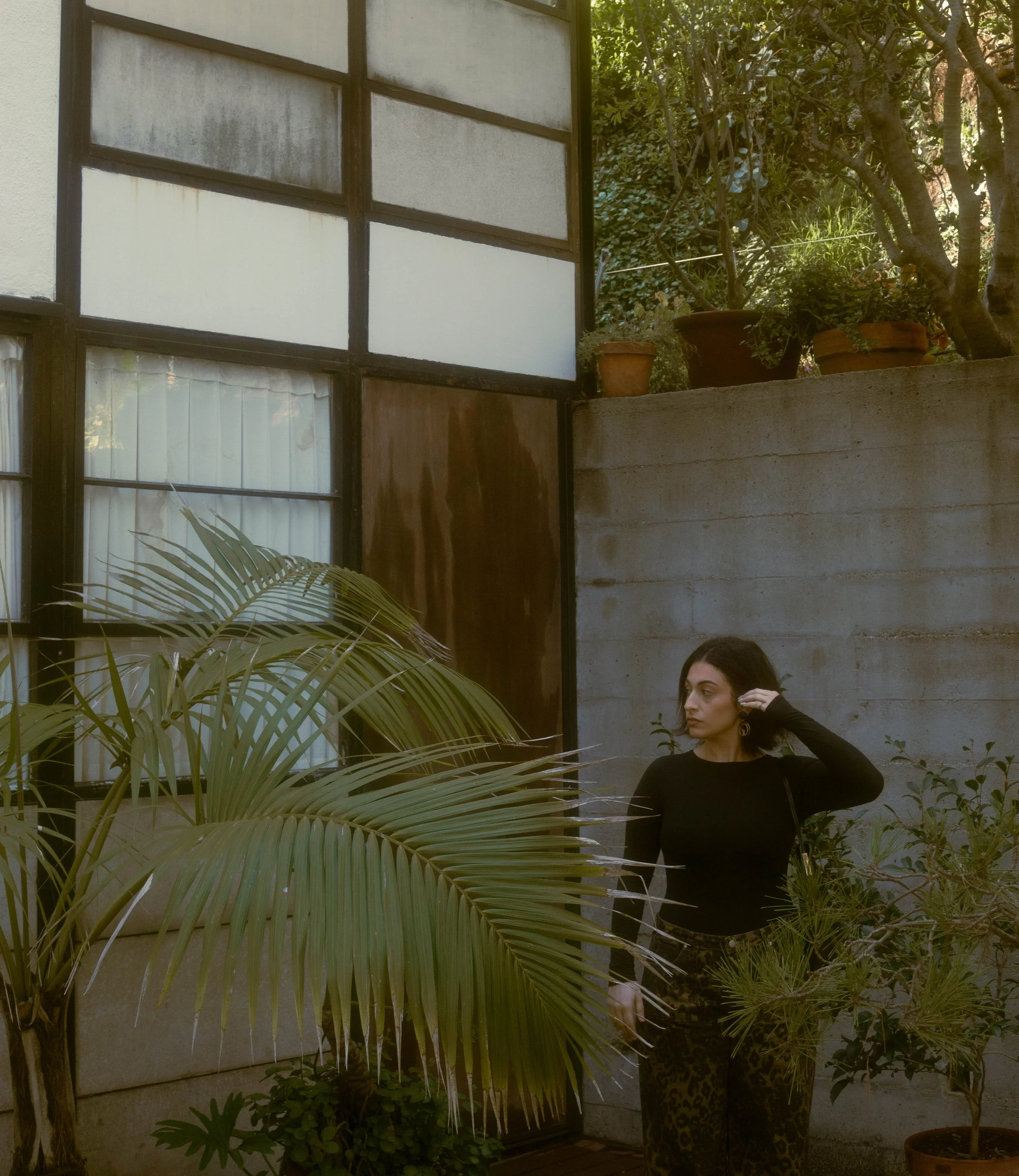 A woman with dark hair and earrings standing outdoors next to a potted plant and green foliage, with a concrete wall and glass window in the background.