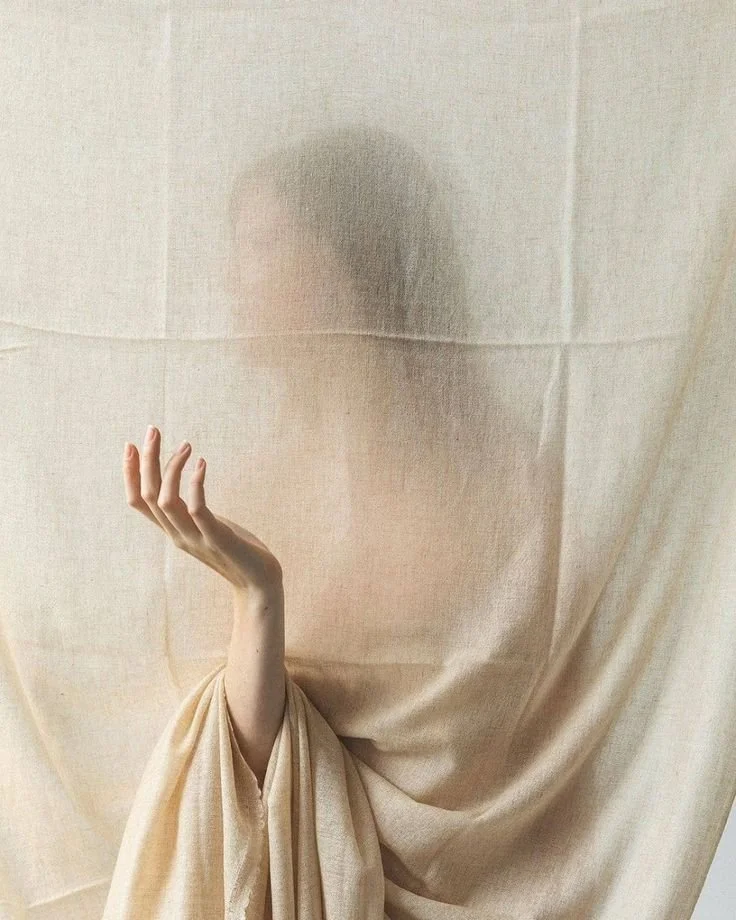 A person behind a semi-transparent fabric, with only their arm and hand visible, holding their fingers in a delicate gesture.