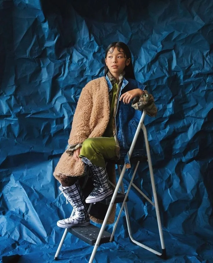 A young woman sitting on a stepladder against a crumpled blue paper background. She wears a tan fuzzy coat, green pants, a plaid shirt, and a denim vest, with checkered high-top sneakers.