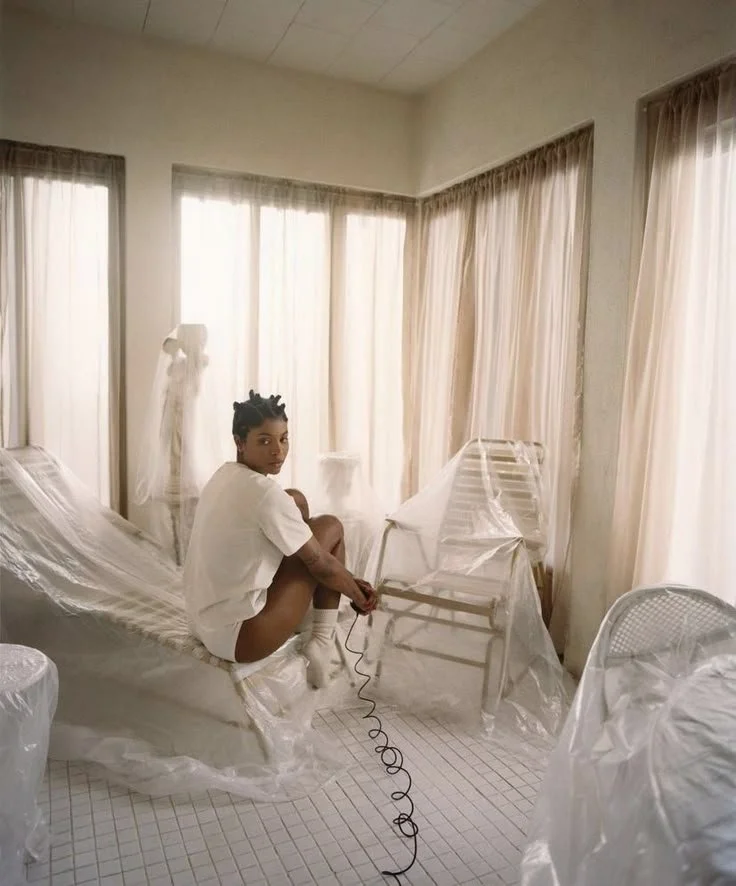 A woman sitting on a bed in a room with sheer curtains, surrounded by plastic-covered furniture and a person holding a phone.
