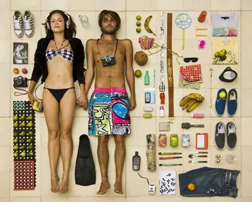 Two young adults lying on a light-colored wooden floor surrounded by summer-related objects, including beach shoes, sunglasses, a camera, fruits, sunscreen, and pool accessories.