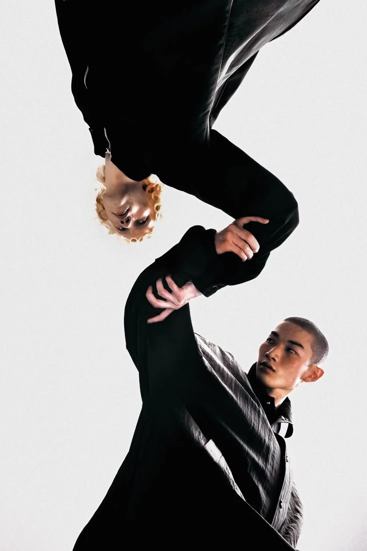 A person with short hair holding another person with curly blonde hair upside down. Both are dressed in black, against a plain white background.