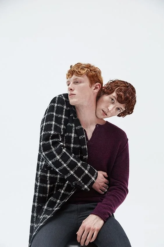 Two young men with red hair, one in a black and white checkered coat and the other in a maroon sweater, sit closely with one leaning on the other's shoulder against a plain white background.