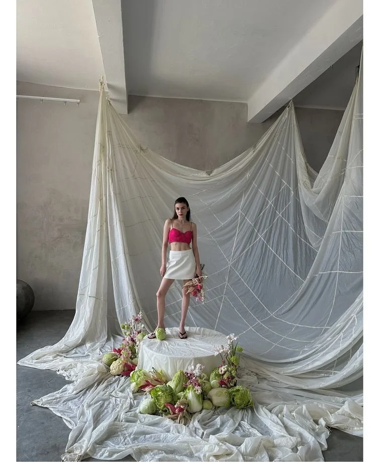 A woman stands on a circular platform surrounded by white and pink flowers with green stems, in front of a backdrop of sheer white fabric draped on a plain wall.