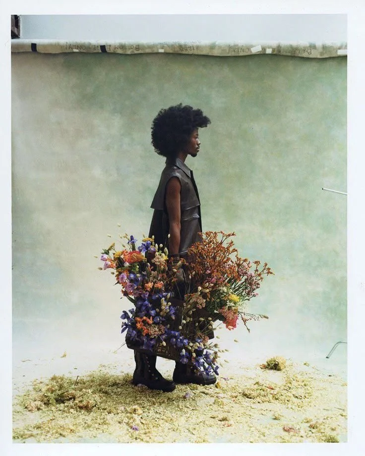 A woman with natural curly hair in profile view, holding a large bouquet of multicolored flowers, standing barefoot on a textured surface against a plain light green background.