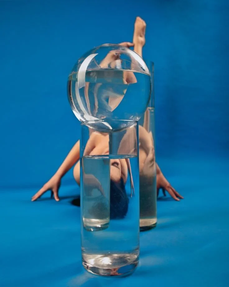 A person performing an acrobatic pose with multiple glass containers and a reflective glass ball, against a blue background.