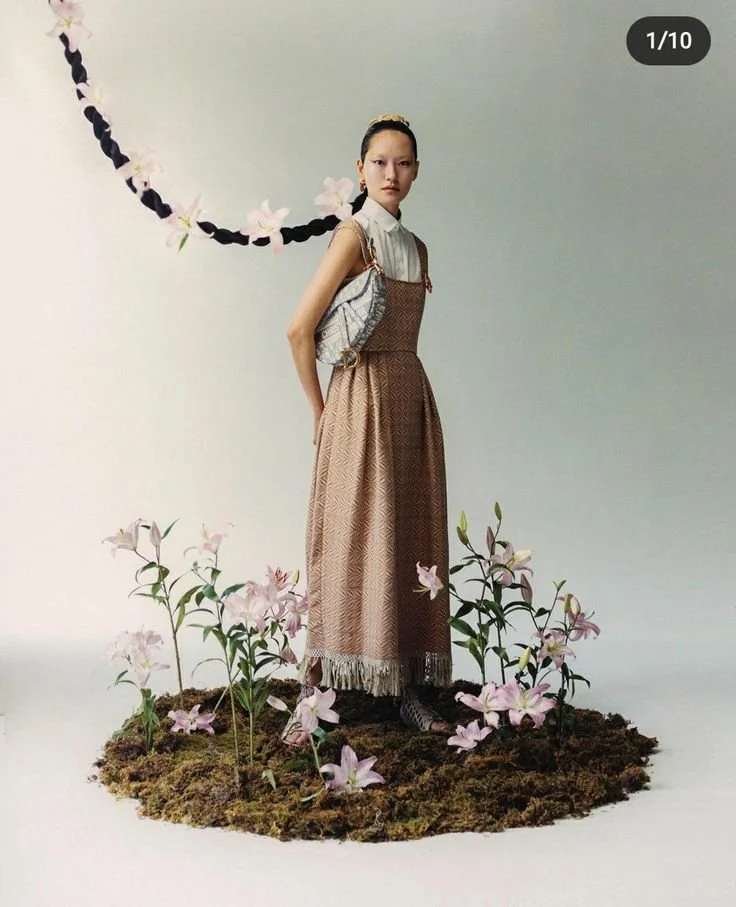 A young woman standing on a patch of moss with pink lilies, against a plain background. She wears a long, patterned dress, a sleeveless top, and has her hair styled with a decorative hairpiece, with a floral chain floating nearby.