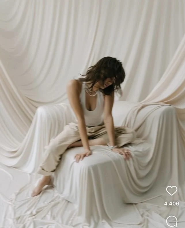 A woman with dark hair wearing a white tank top and beige pants, sitting on a draped white fabric-covered surface, with draped white fabric background.