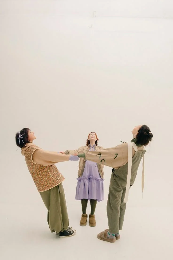 Three women holding hands and leaning back while standing in a white space, smiling and looking up.