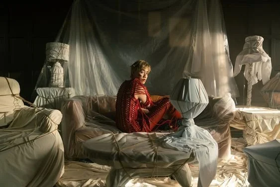 A woman sitting on a beige sofa in a dimly lit room with draped sheets and white bust sculptures, wearing a red outfit and looking at the camera.