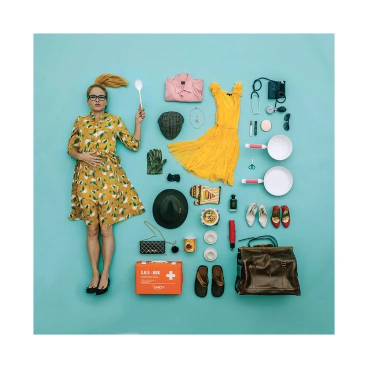 A woman lying on a blue background surrounded by various travel and fashion items, including a yellow dress, pink shirt, black hat, purse, shoes, camera, and an orange first aid kit.