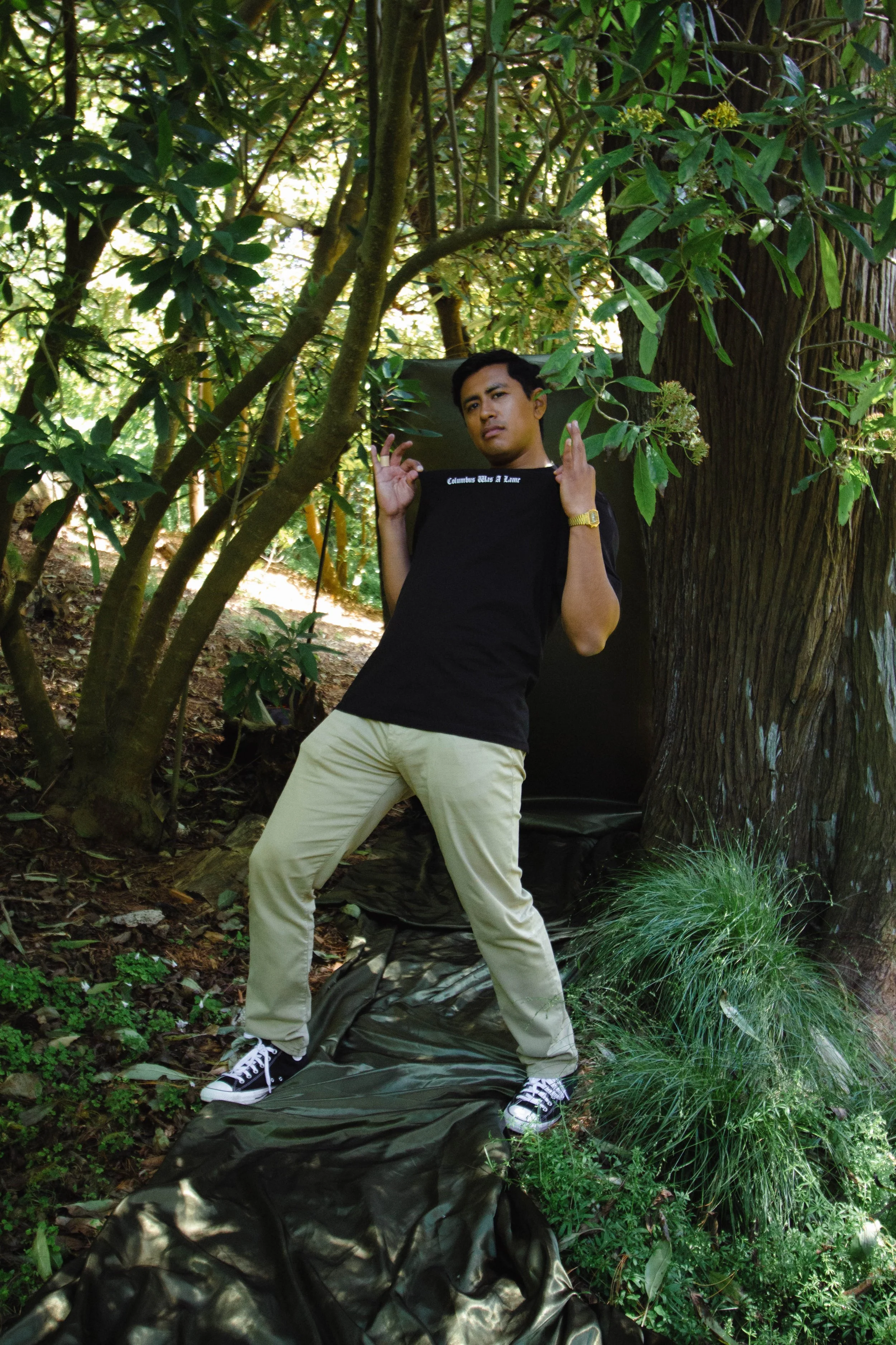 A young man stands outdoors amid green foliage, leaning against a large tree trunk, wearing a black t-shirt, beige pants, black sneakers, and a gold watch. He is posing with both hands raised near his face, looking serious.