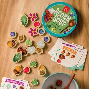 A board game called "Acorn Soup" featuring small wooden food tokens like trees, flowers, acorns, berries, and ice cream cones, along with recipe cards, a bowl, and a spoon on a wooden surface.