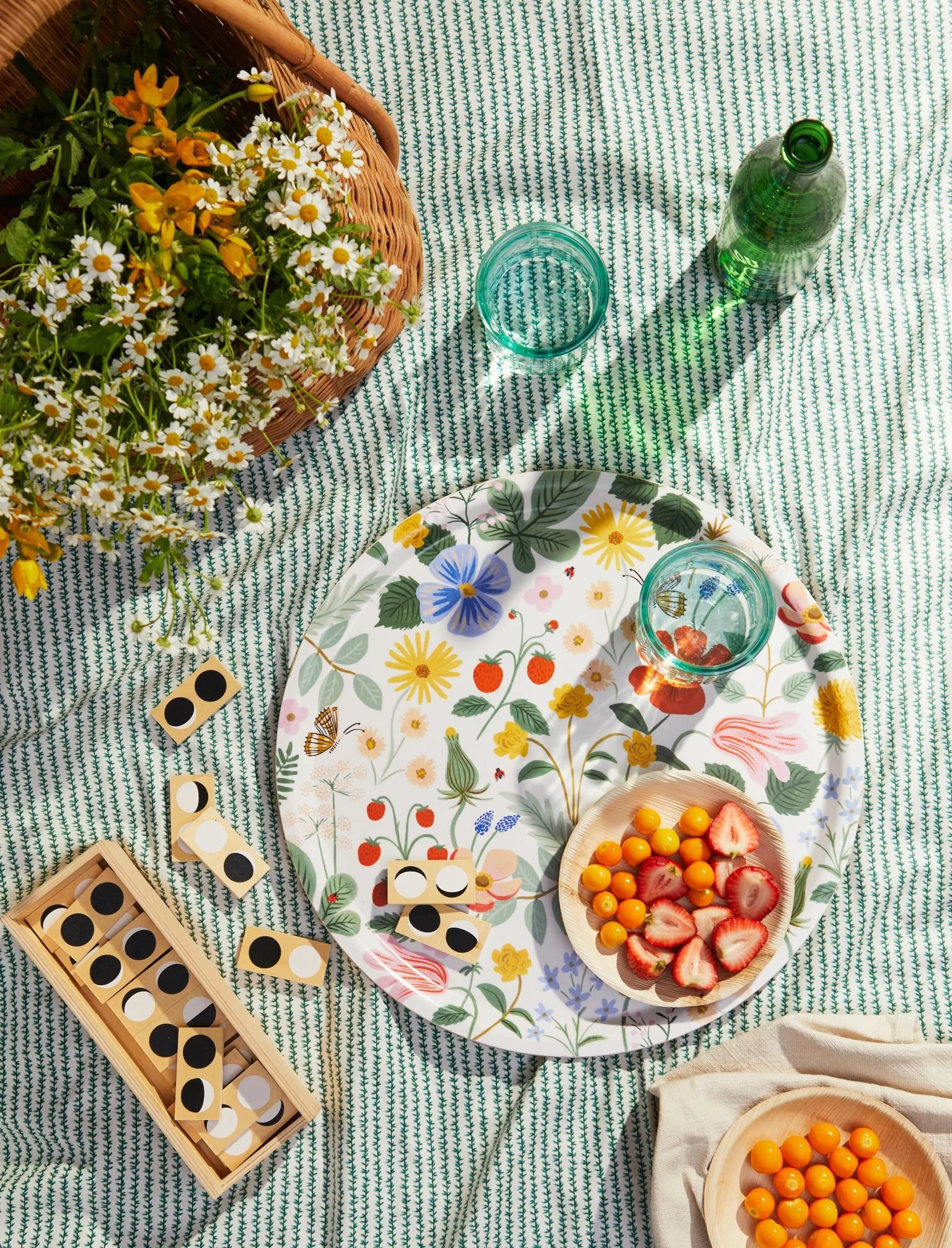 A picnic setup on a green patterned tablecloth featuring a basket of daisies and yellow flowers, a large floral platter with strawberries and cherry tomatoes, wooden dominoes, and drinking glasses, with small bowls of cherry tomatoes and strawberries.