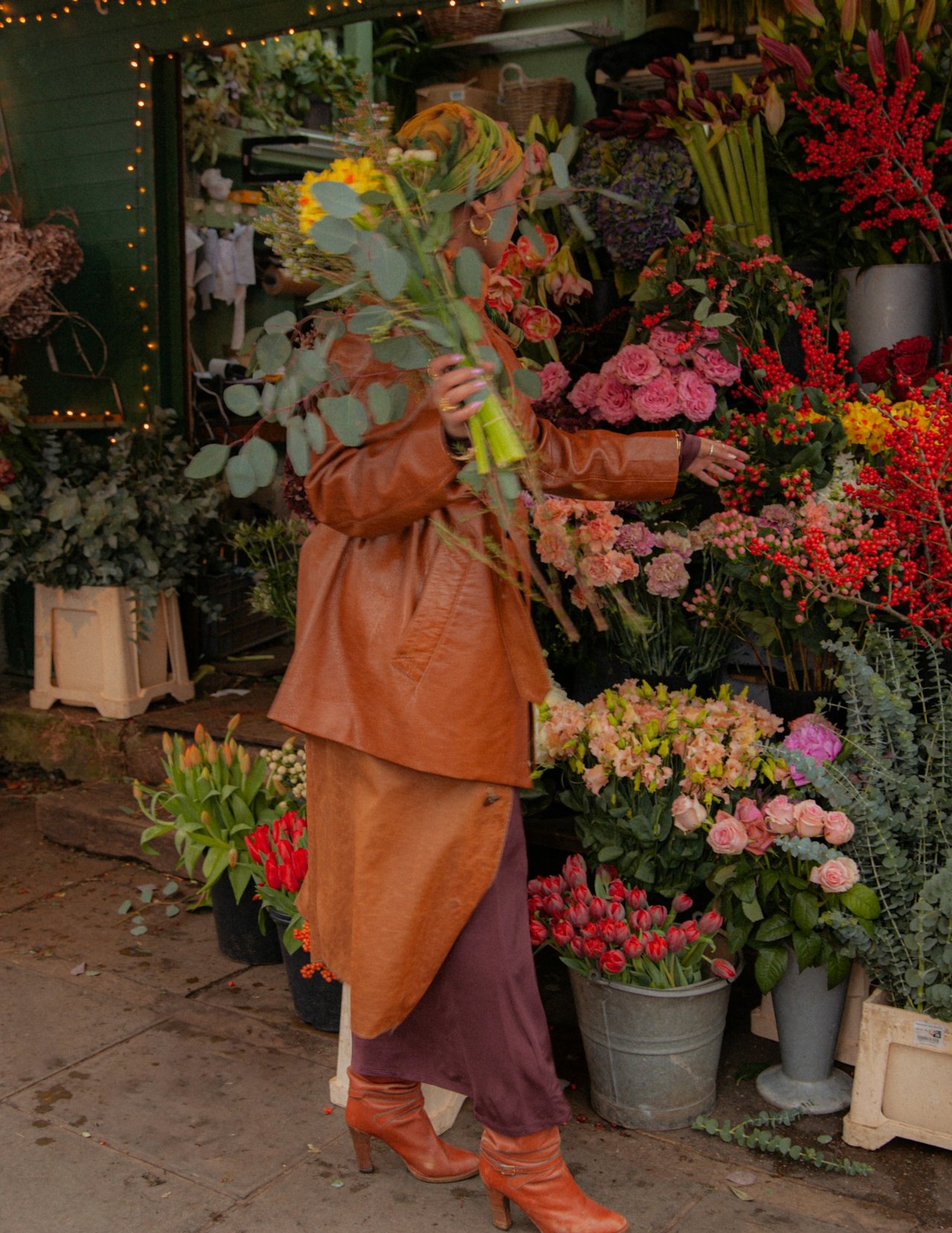 A woman shopping for flowers at an outdoor flower stand, holding a bouquet of yellow flowers. She is dressed in a brown leather coat, brown boots, and a colorful headscarf, surrounded by various colorful flowers including tulips, roses, and other blossoms.